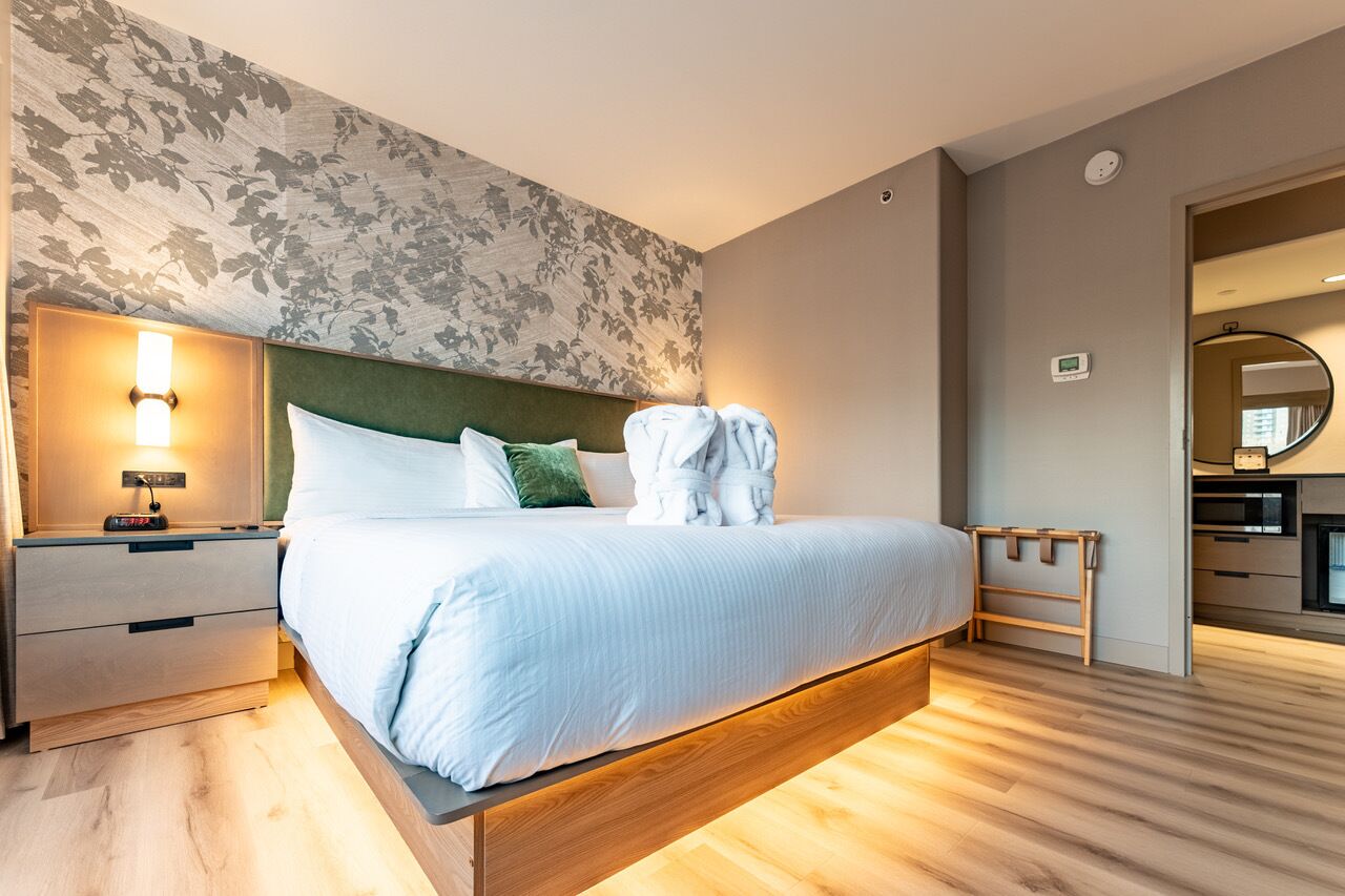 Una habitación de hotel moderna con una cama grande, una mesita de noche, un banco de madera y vistas al área de la cocina.