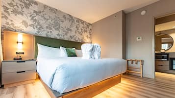 Una habitación de hotel moderna con una cama grande, una mesita de noche, un banco de madera y vistas al área de la cocina.