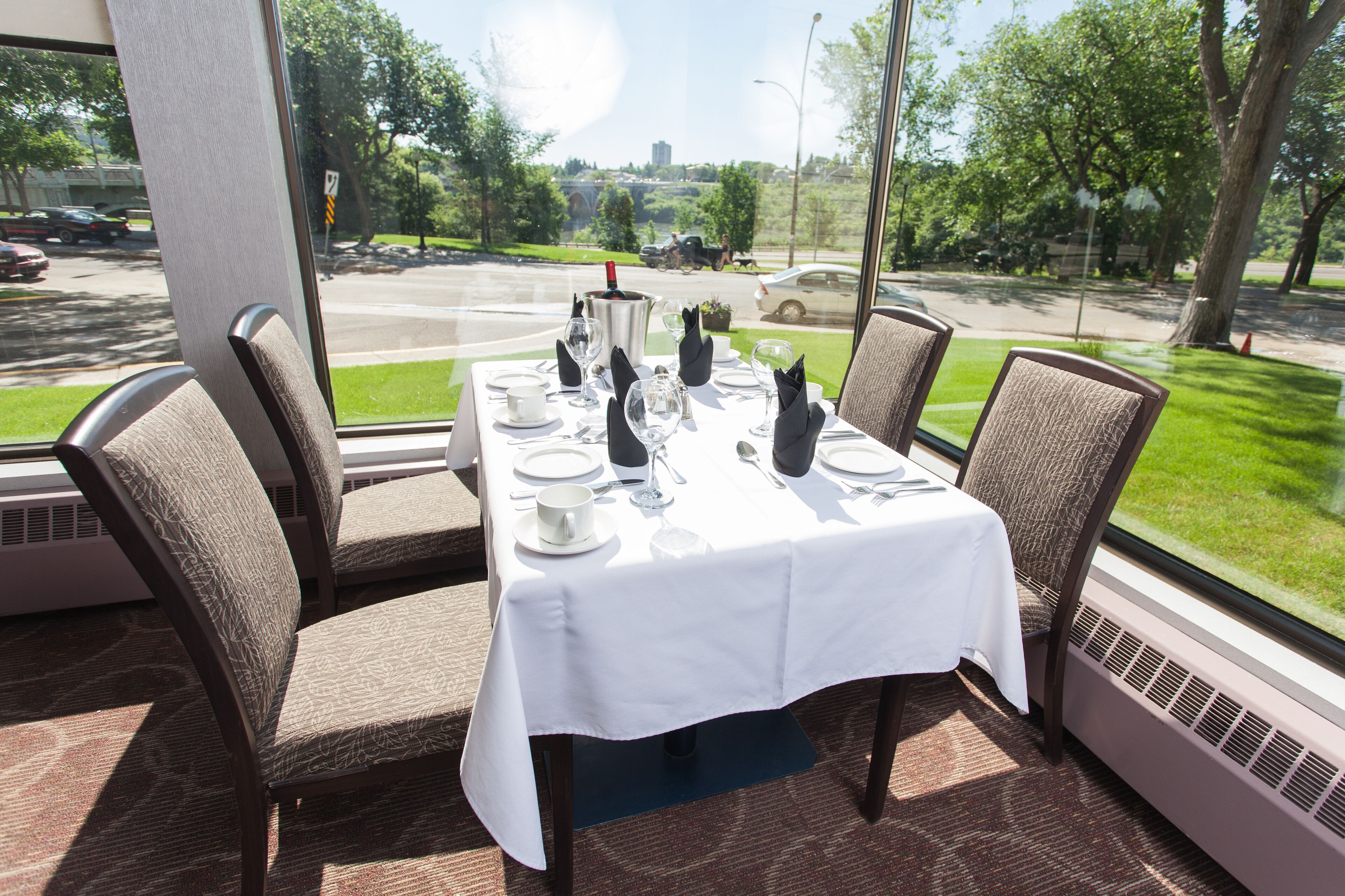 Mesa de comedor preparada para cuatro personas, con mantel blanco, cuatro sillas y vista a una calle con coches y áreas verdes.