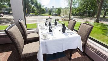 Mesa de comedor preparada para cuatro personas, con mantel blanco, cuatro sillas y vista a una calle con coches y áreas verdes.
