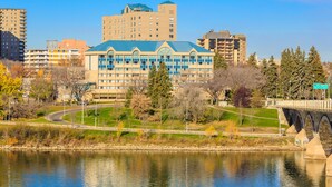 A large building with a blue roof and multiple windows, surrounded by trees and a body of water. - Park Town Hotel (Saskatoon)