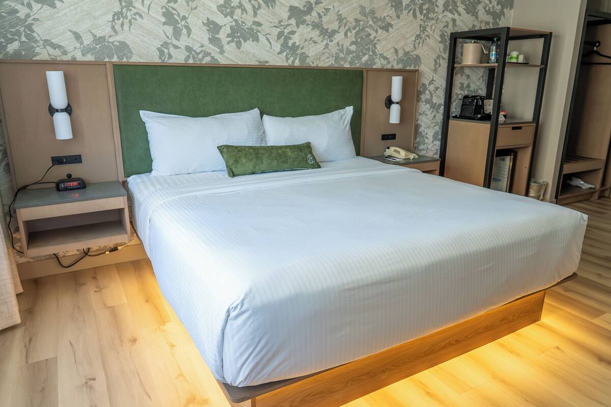 A hotel room with a large bed, wooden headboard, bedside tables, a nightstand, and a built-in shelf with a kettle and coffee maker.