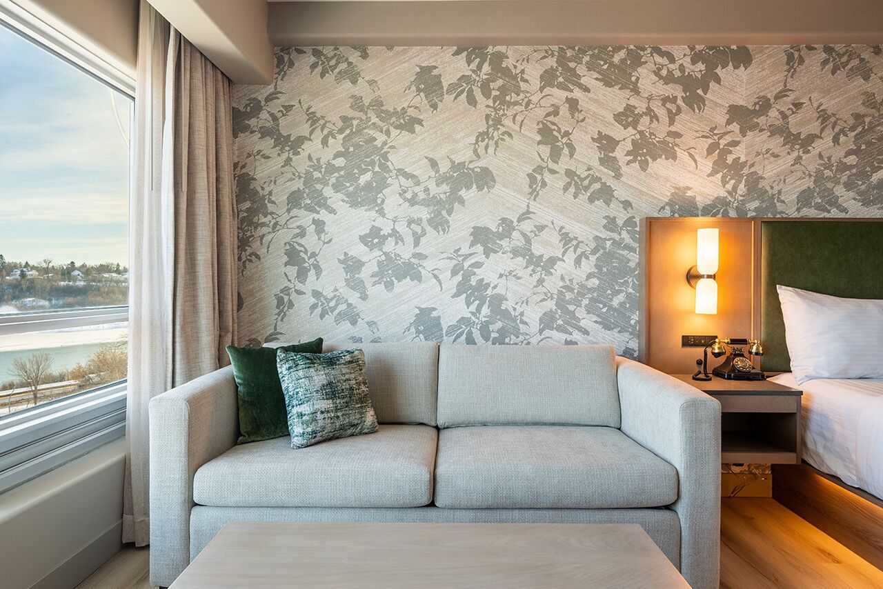 A hotel room with a sofa, a bedside table with a lamp, a bed, and a window with curtains.