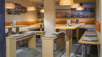 A modern, well-lit restaurant with wooden tables and benches, overhead woven light fixtures, and a wall adorned with framed photos and a large mural.