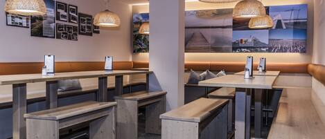 Un restaurante moderno con mesas y bancos de madera, lámparas colgantes tejidas y una pared decorada con diversas fotografías.