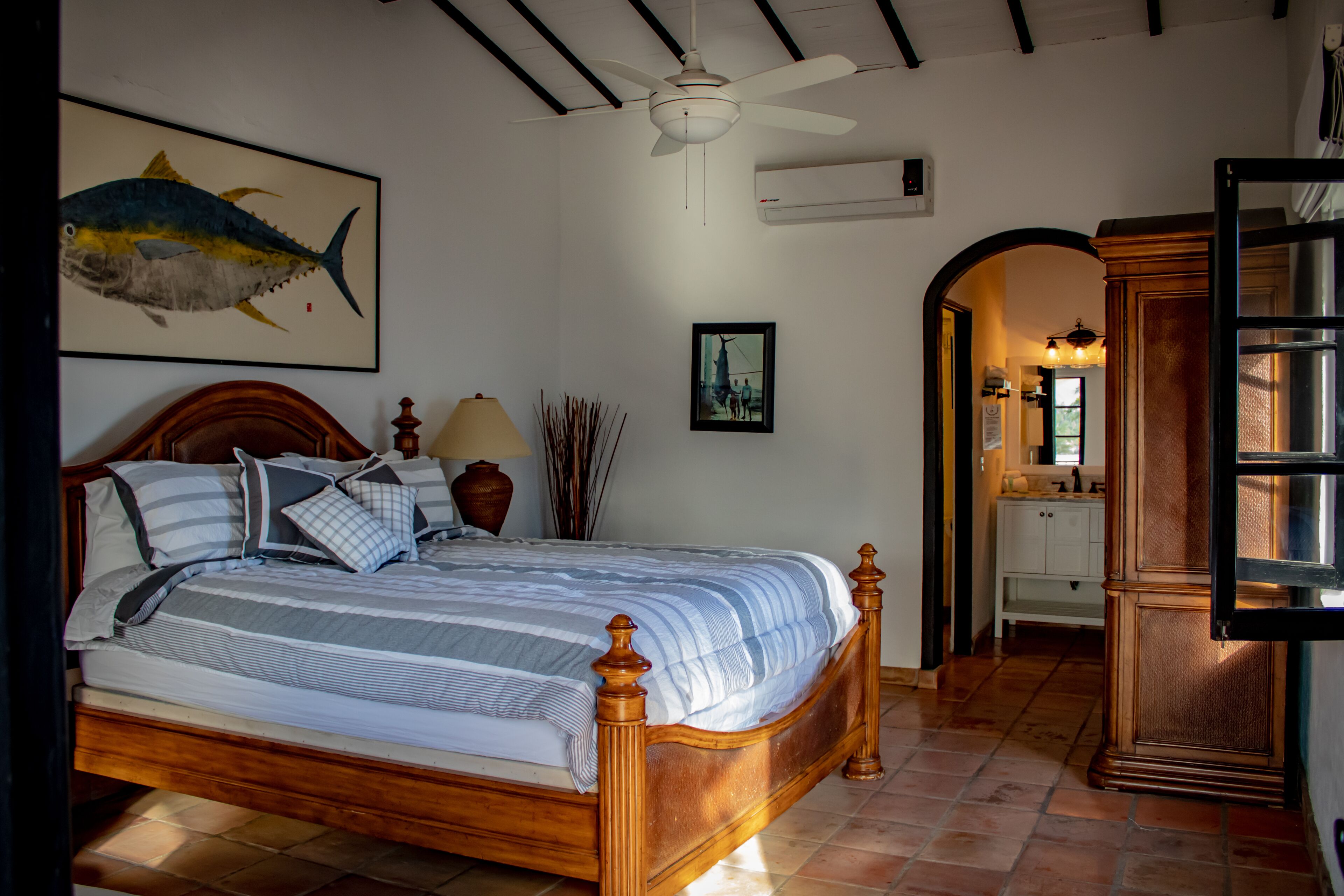 A bedroom with a wooden bed, a ceiling fan, a framed fish painting, and a view of the bathroom.