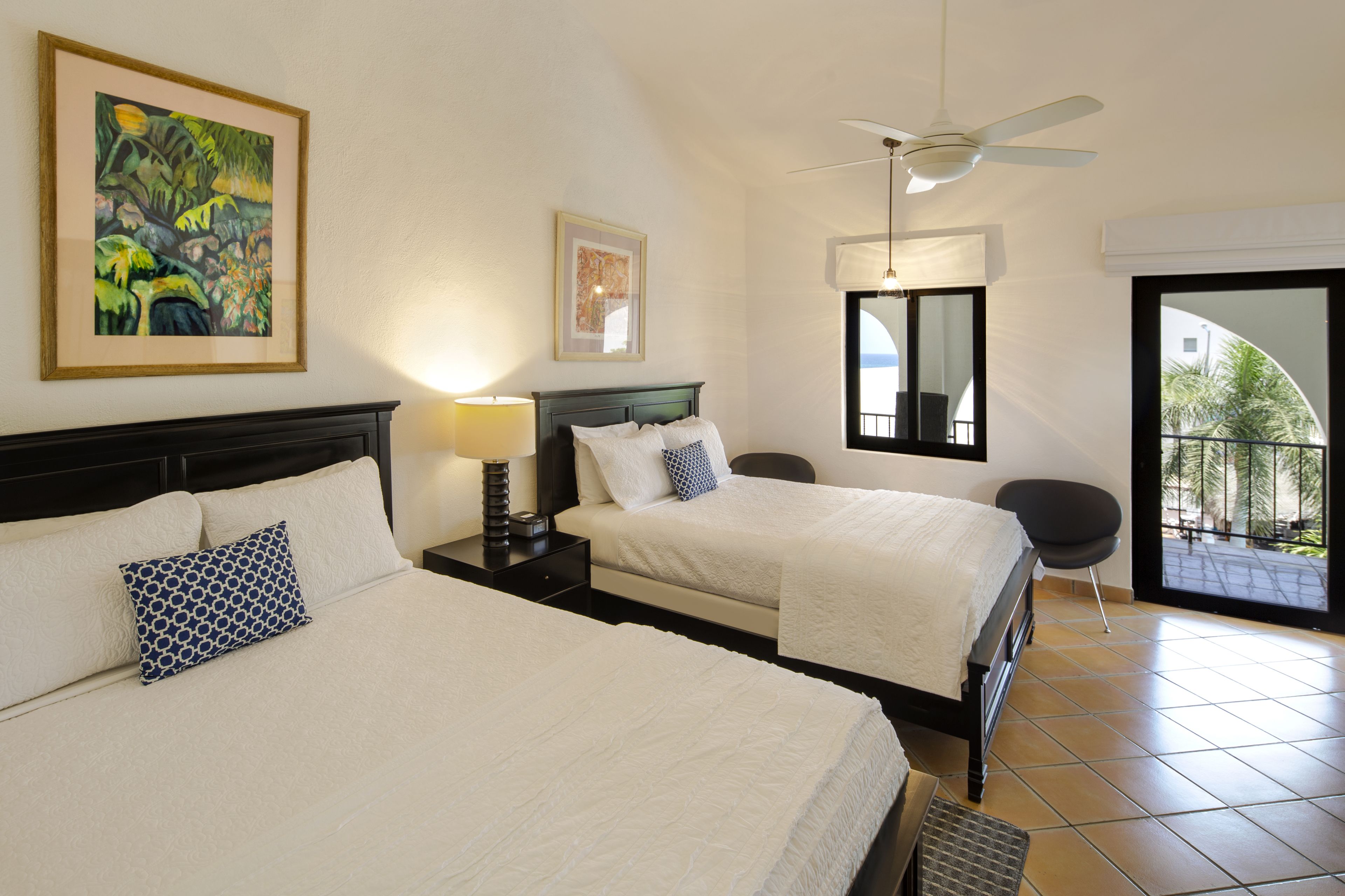 Habitación de hotel con dos camas, ventilador de techo, un ventanal grande y obras de arte en las paredes.