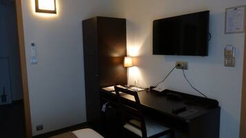 A small hotel room with a desk, chair, lamp, television, and a wall-mounted thermostat.