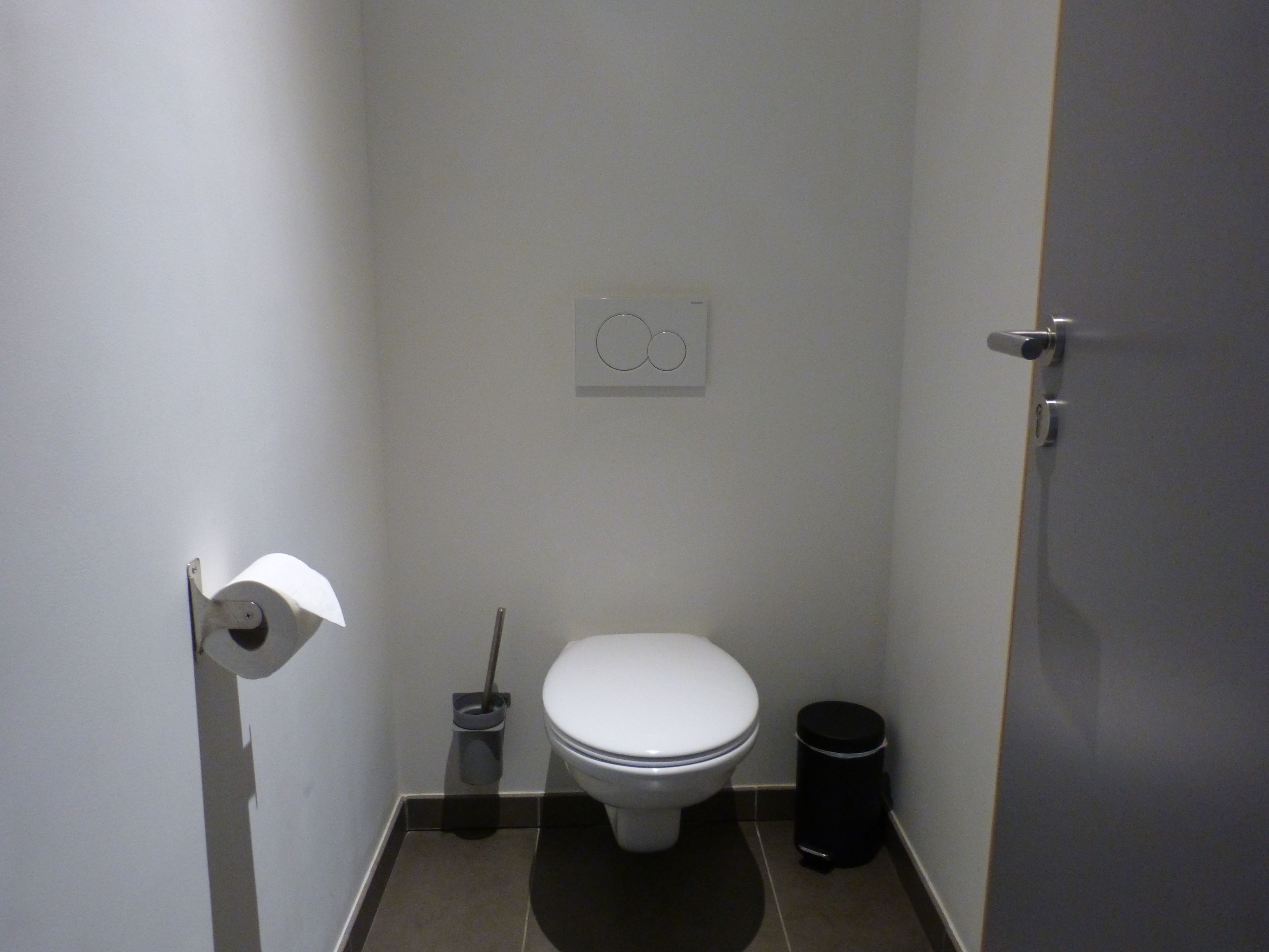 Een kleine, praktische badkamer met een toilet, een prullenbak en een wc-papierhouder.