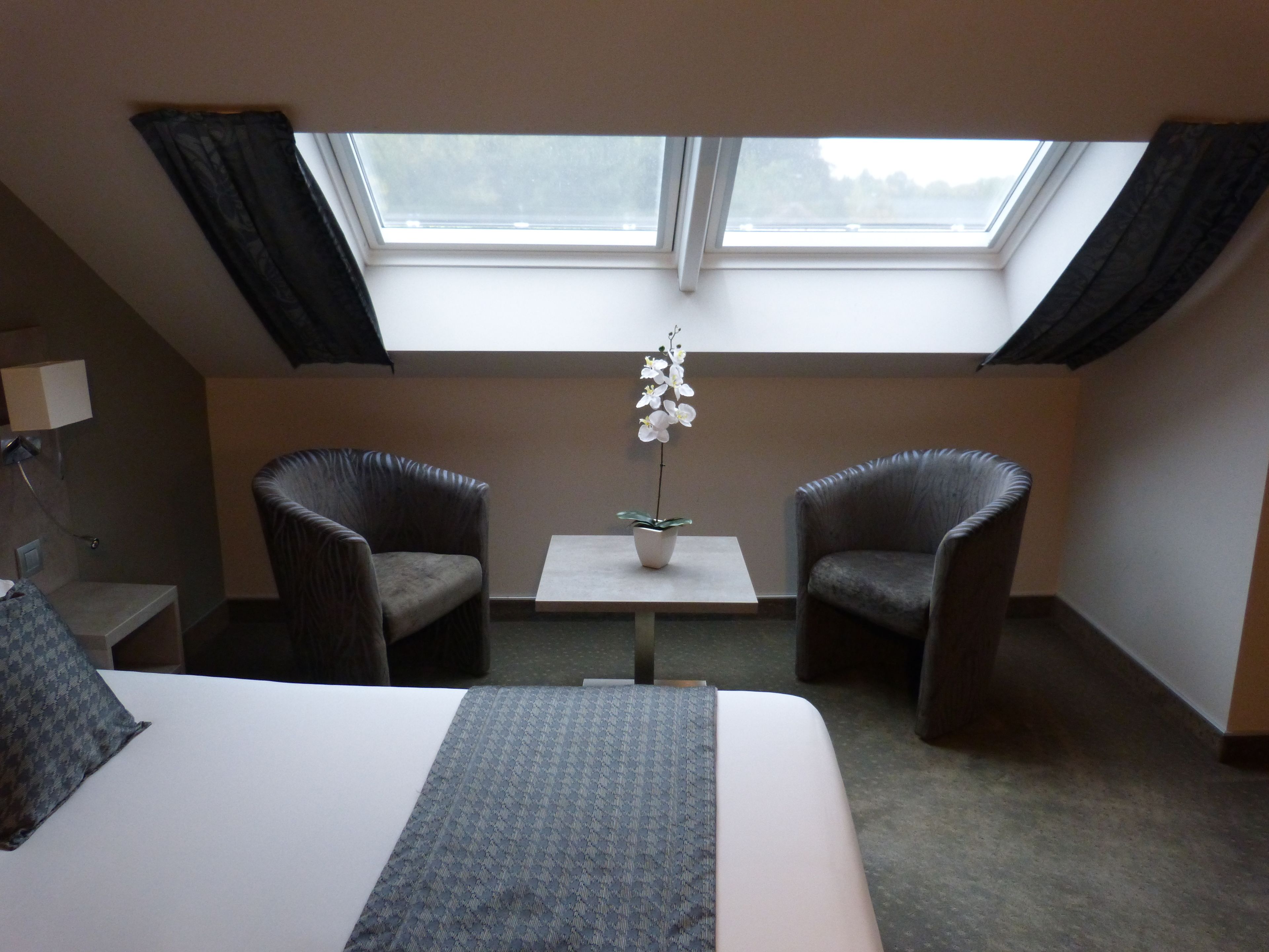Habitación de hotel con cama, dos sillones, una mesita con una orquídea en maceta y un tragaluz.