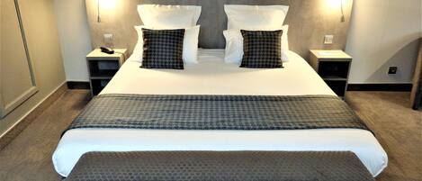Una cama bien tendida con una colcha gris y blanca a cuadros, dos mesitas de noche con lámparas y un banco gris al pie de la cama.