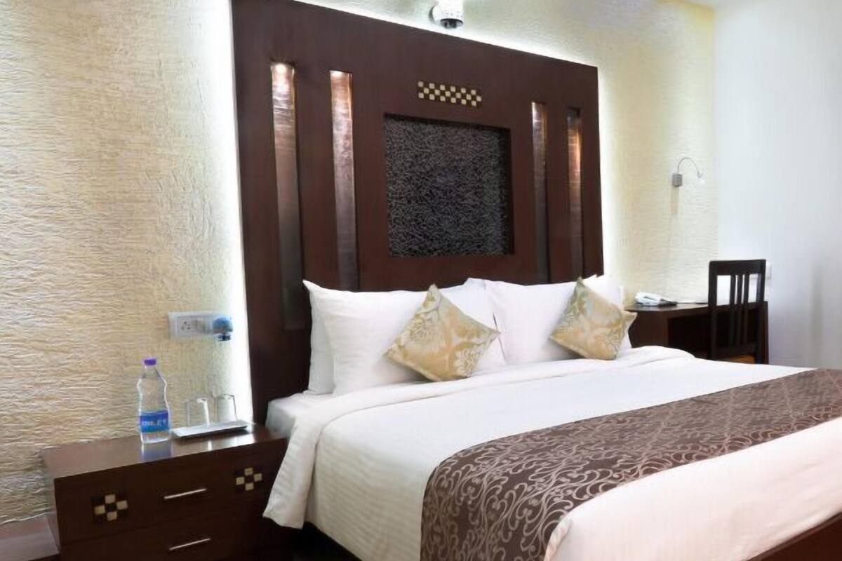 A hotel room with a large bed, a wooden nightstand, a small table, and a wall-mounted light fixture.
