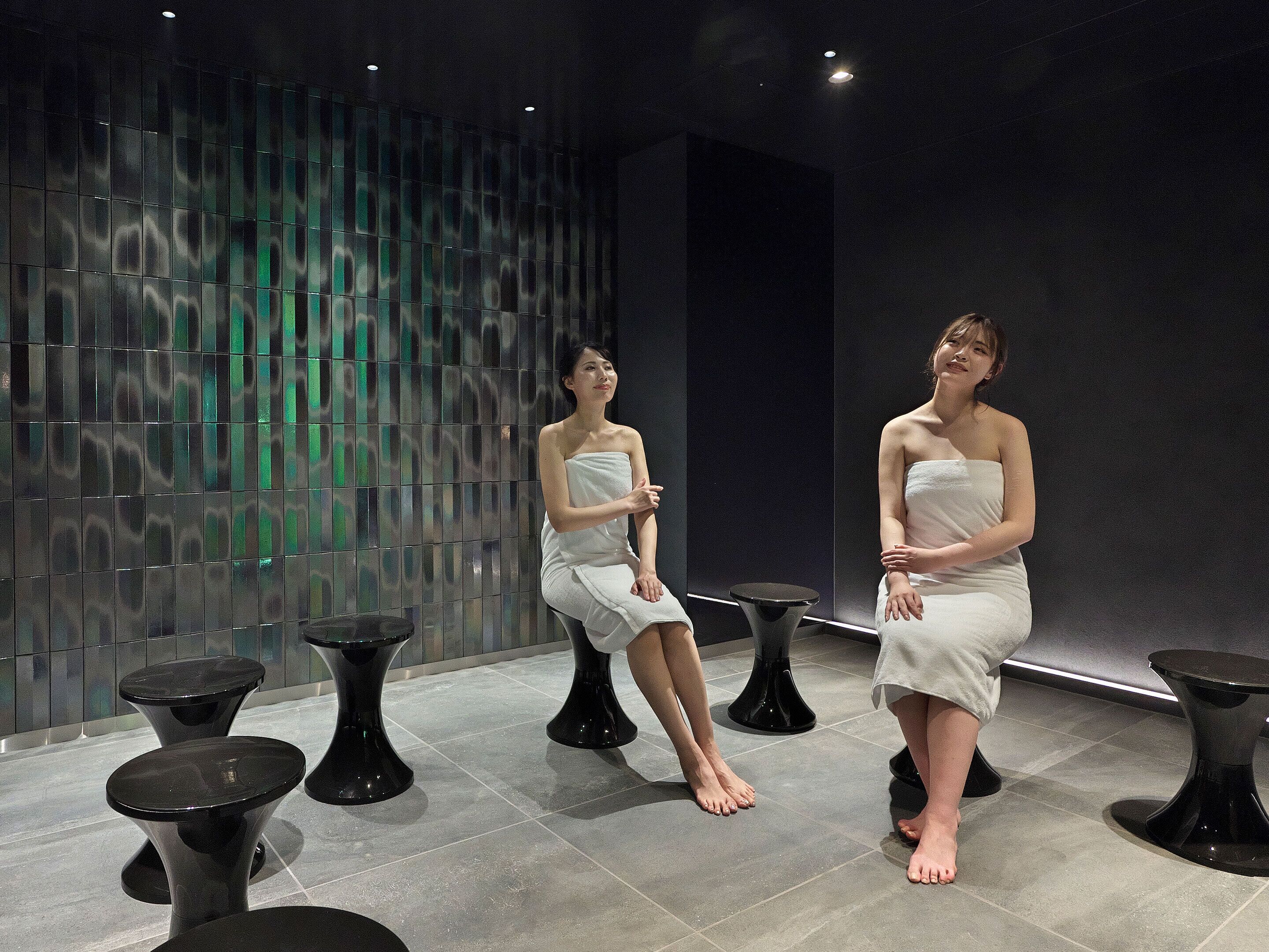 A modern spa room with two women seated on stools, a reflective wall, and black tables.