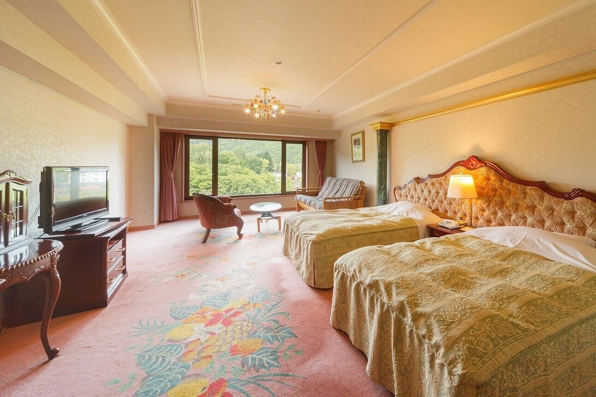 A hotel room with two beds, a television, a desk, and a view of greenery.
