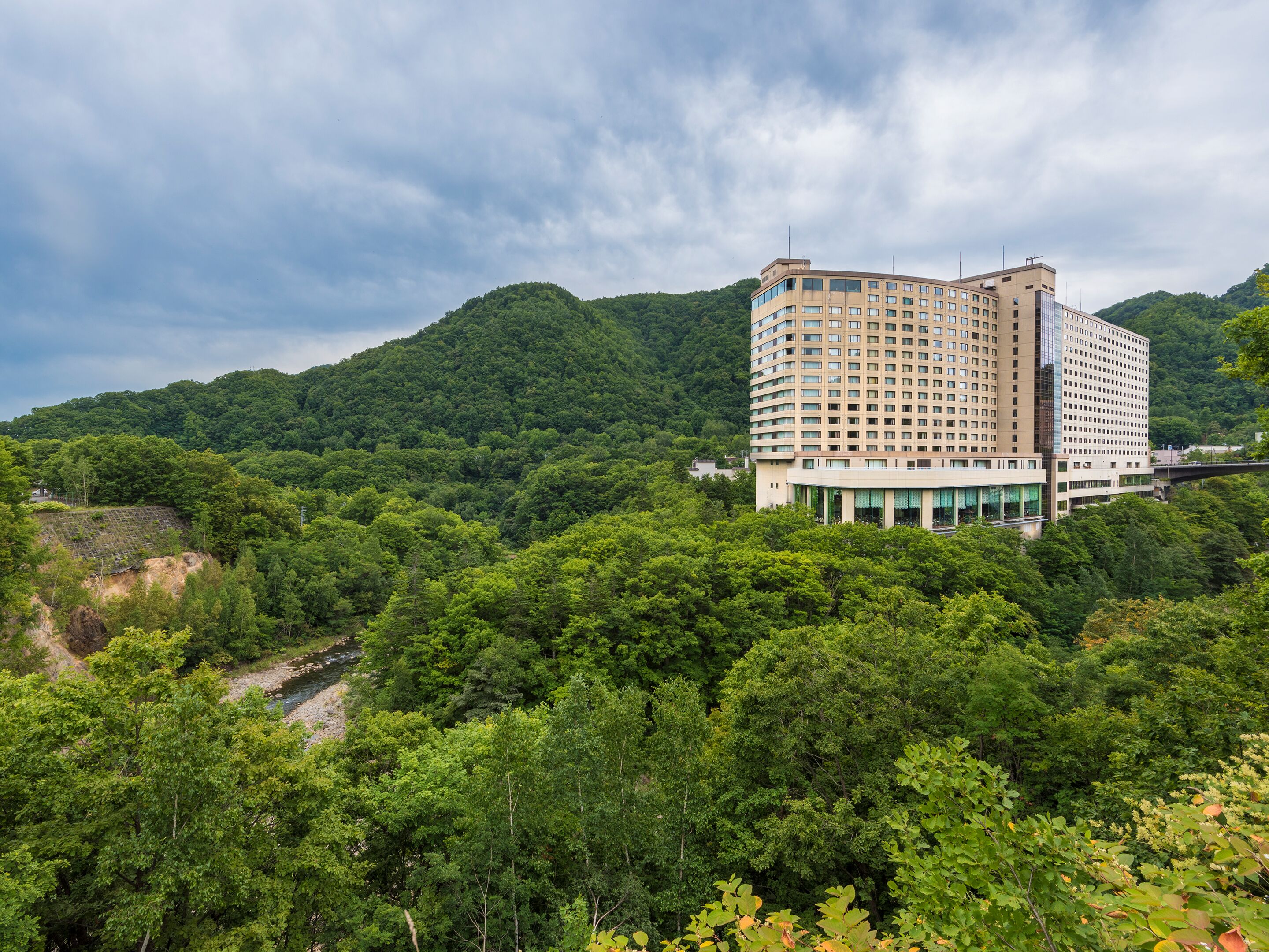 A multi-story hotel with a modern design, surrounded by lush greenery and hills.
