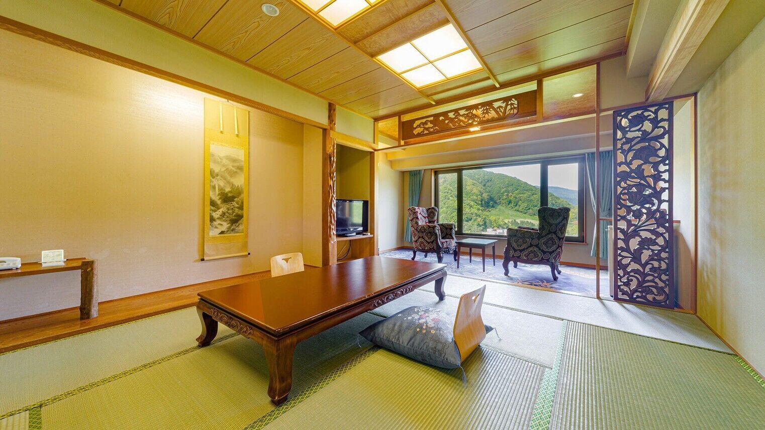 A traditional Japanese-style room with tatami flooring, a low wooden table, a television, and a view of a mountainous landscape through large windows.