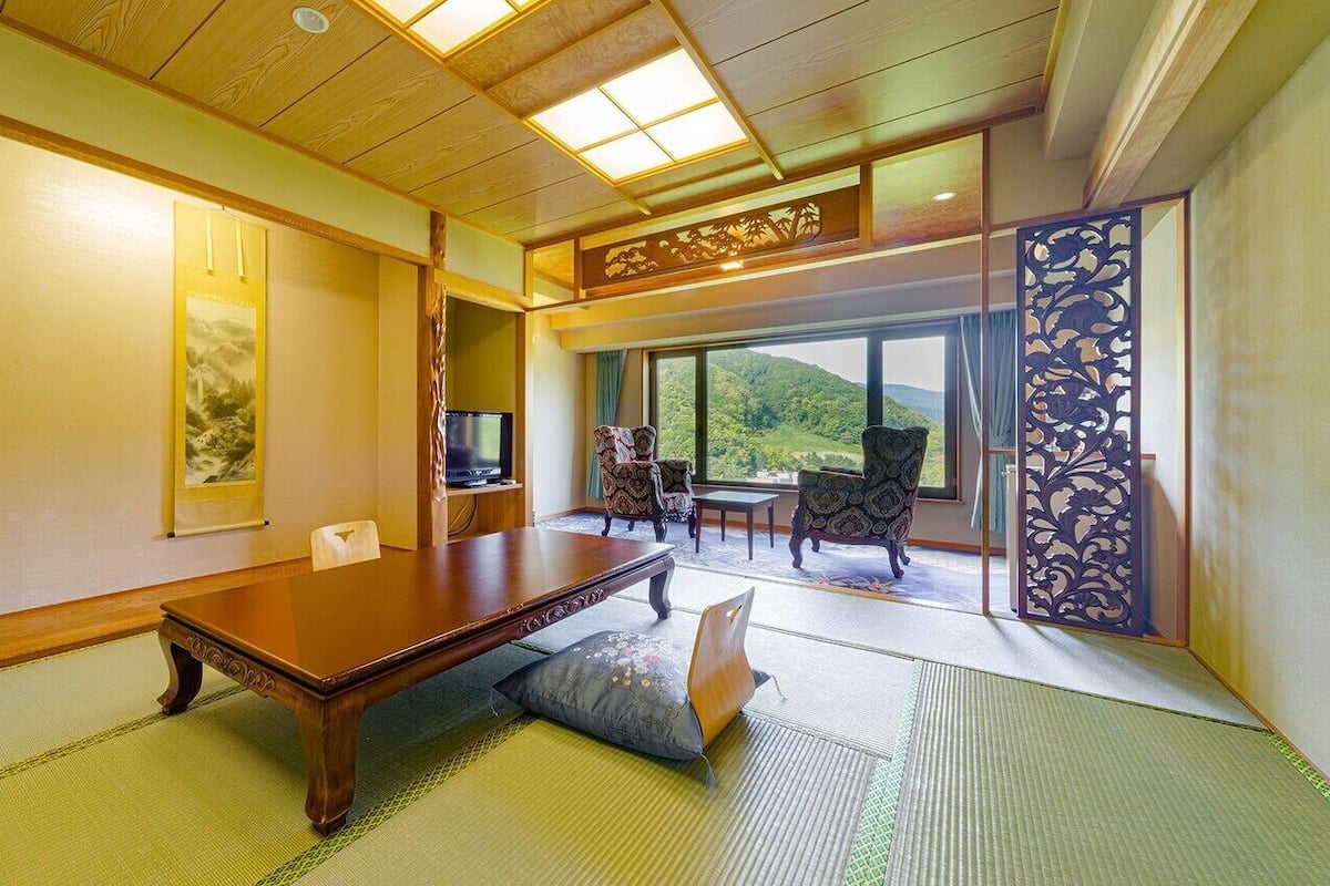 A traditional Japanese-style room with tatami flooring, a low wooden table, a television, and a view of a mountainous landscape through large windows.