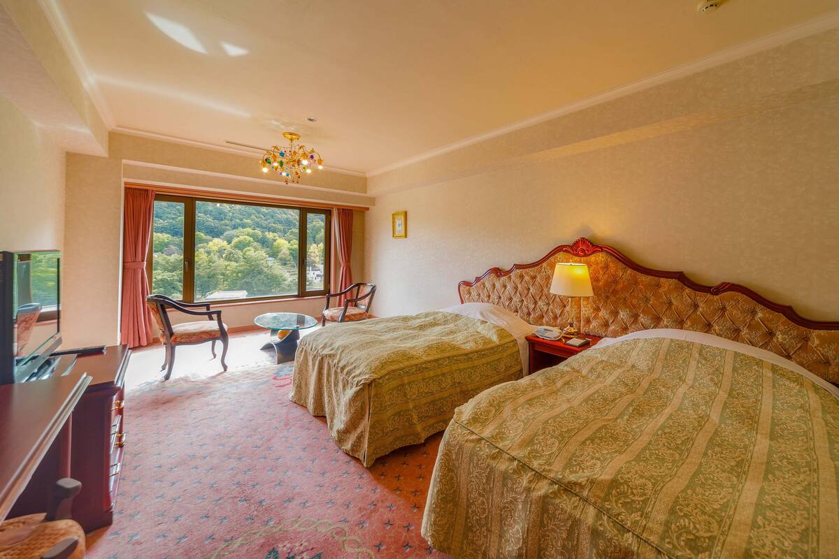 Quarto de hotel com duas camas, uma escrivaninha, uma cadeira, uma mesinha e uma janela grande com vista para o exterior.