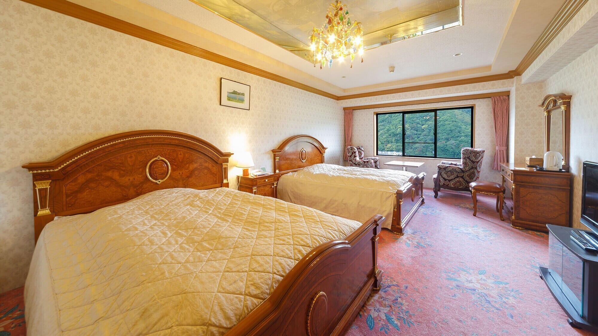 A hotel room with two beds, a television, a desk, and a window with a view of trees.