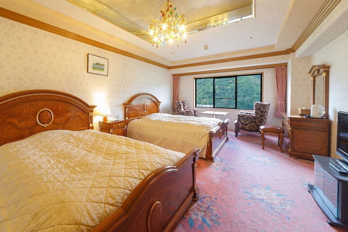 A hotel room with two beds, a television, a desk, and a window with a view of trees.