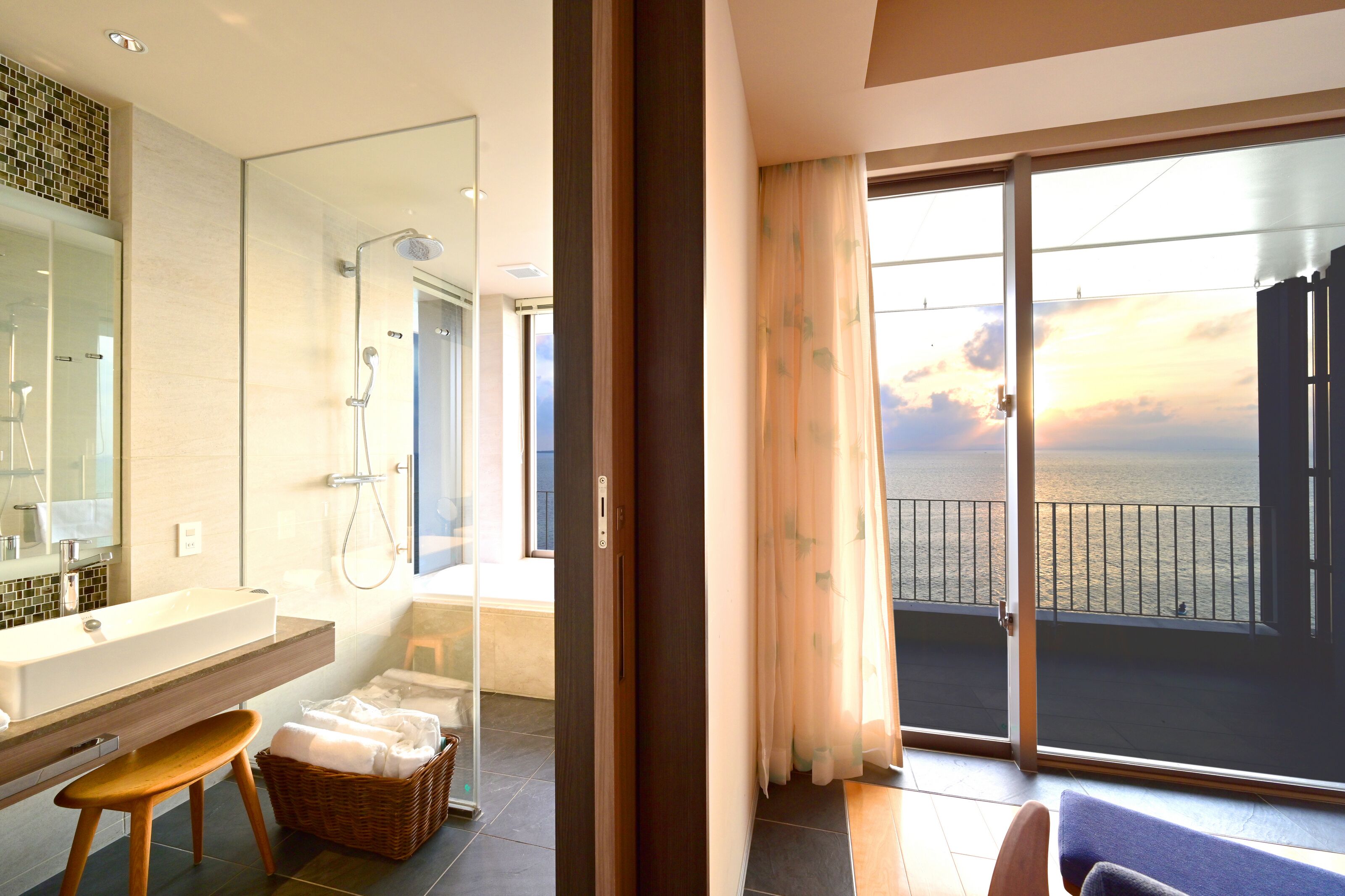 A modern bathroom with a large mirror, a sink, and a shower. There is a balcony with a view of the sea.