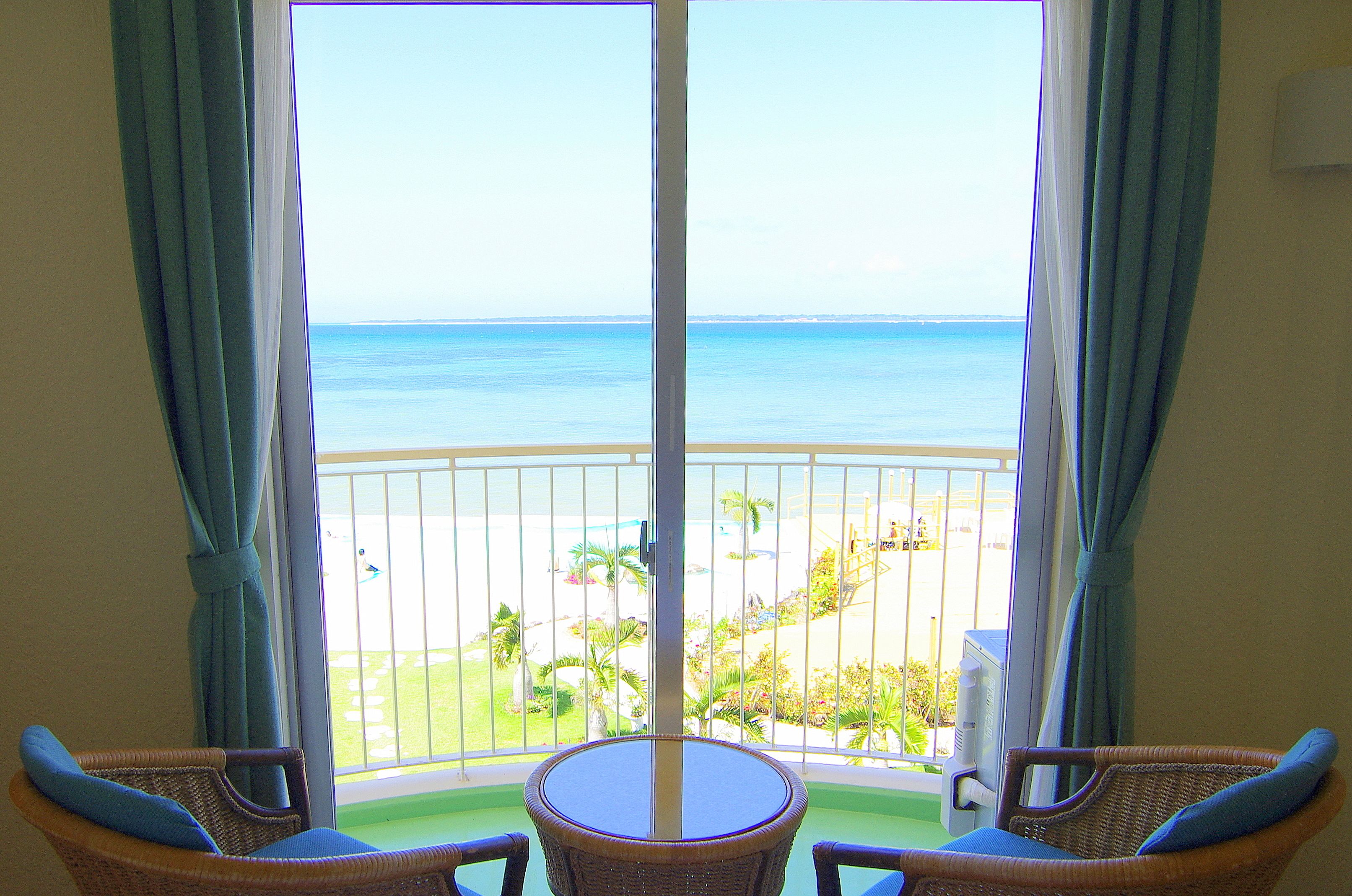Un balcón con sillas de mimbre y una mesa, con vista a una playa con palmeras y una vista despejada del océano.