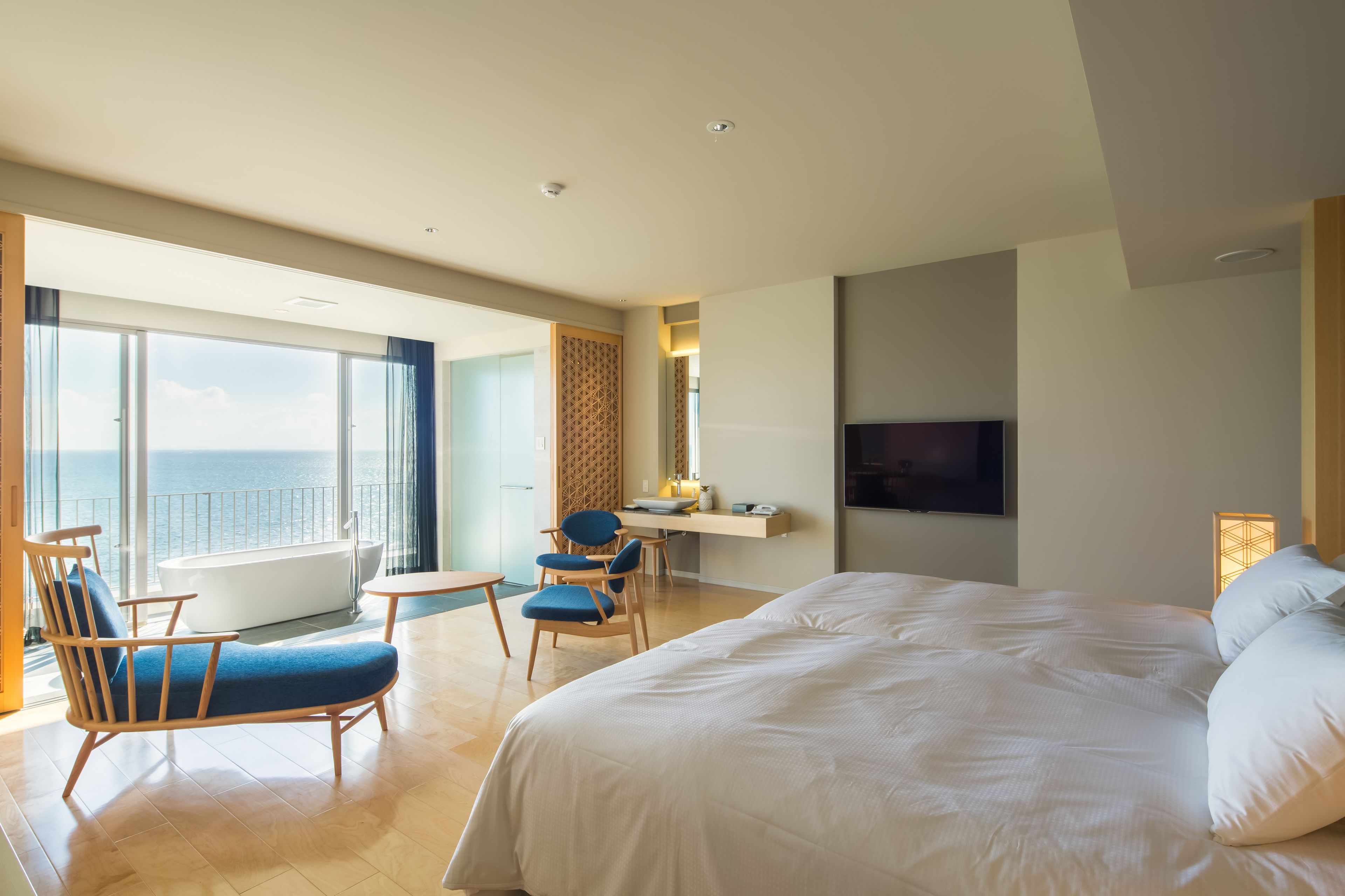 A modern hotel room with a large bed, a desk, a chair, and a view of the sea.