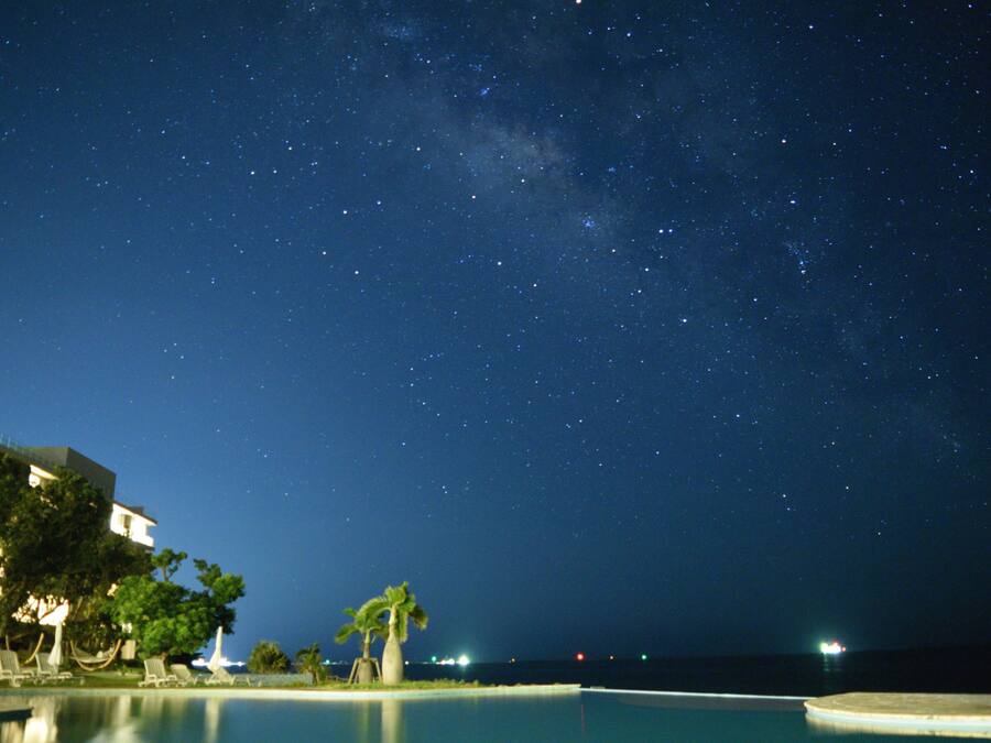Une vue nocturne d’un complexe hôtelier avec une piscine, des palmiers et un ciel étoilé.