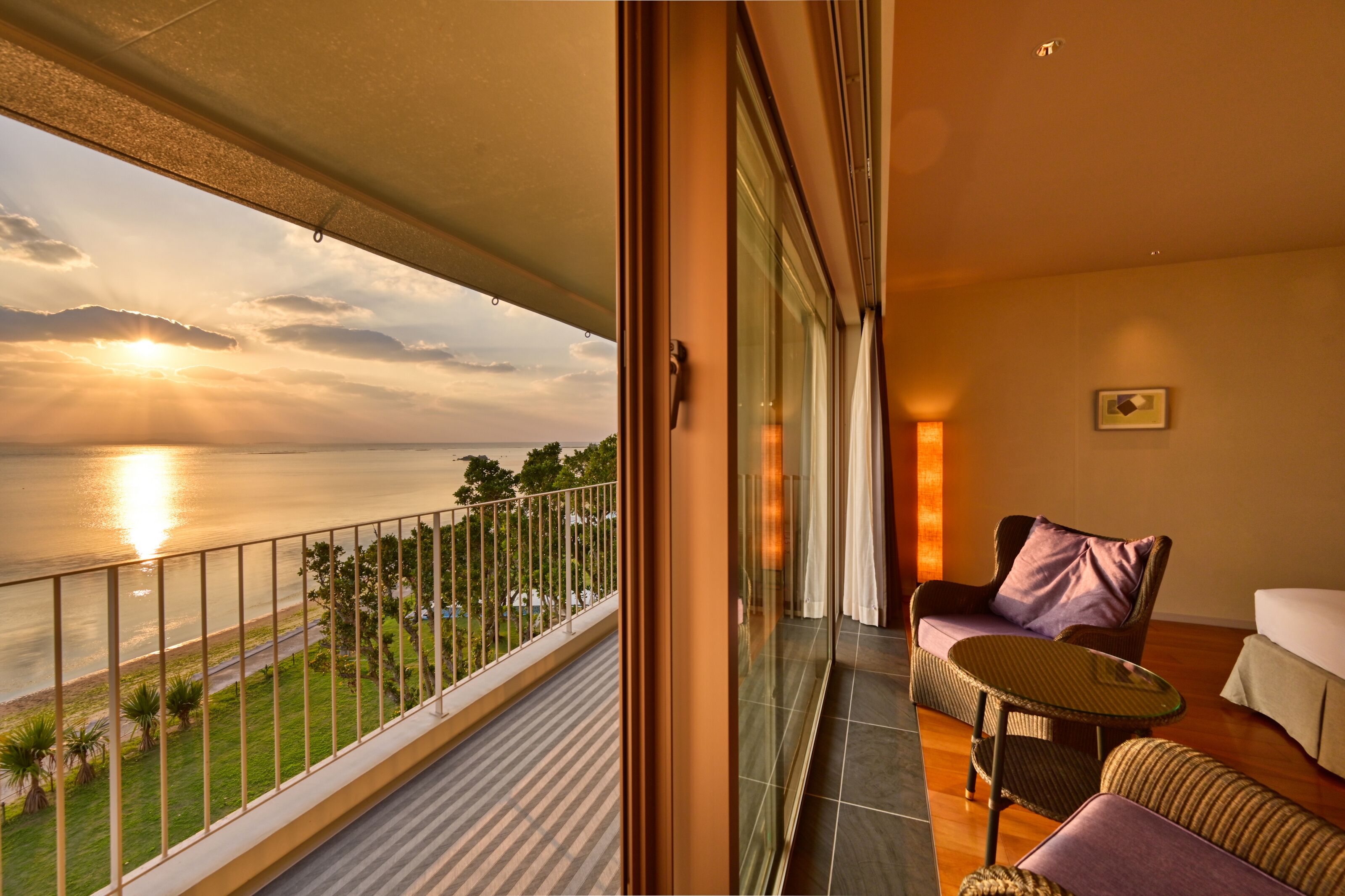 Ein Balkon mit Blick auf den Sonnenuntergang über dem Wasser, eine Rattan-Sofa, ein kleiner Tisch und ein weißes Sofa.