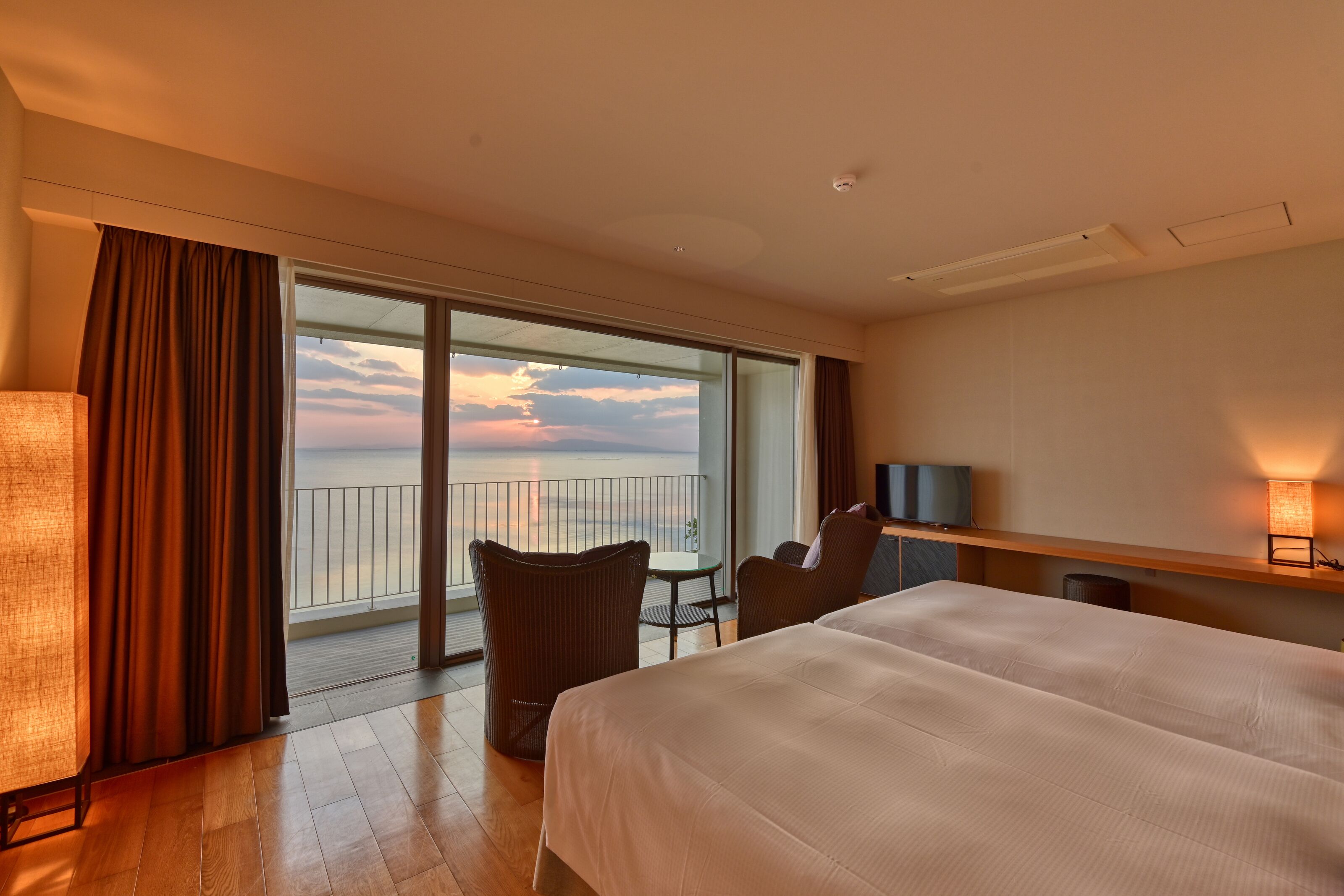A hotel room with a large bed, a desk, a chair, a TV, and a balcony with a view of the sunset.