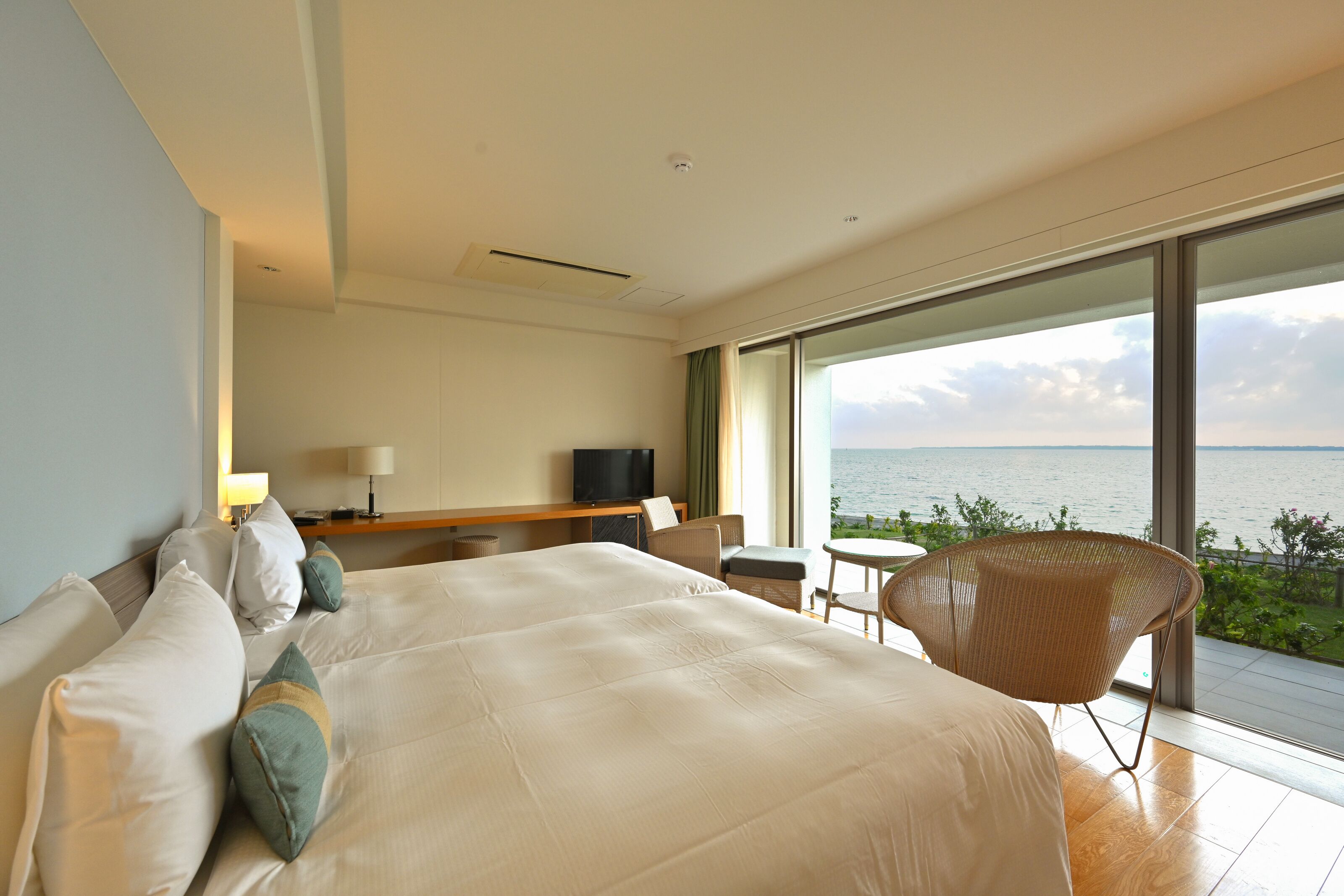A modern hotel room with a large bed, a desk with a TV, a chair, and a view of the sea.
