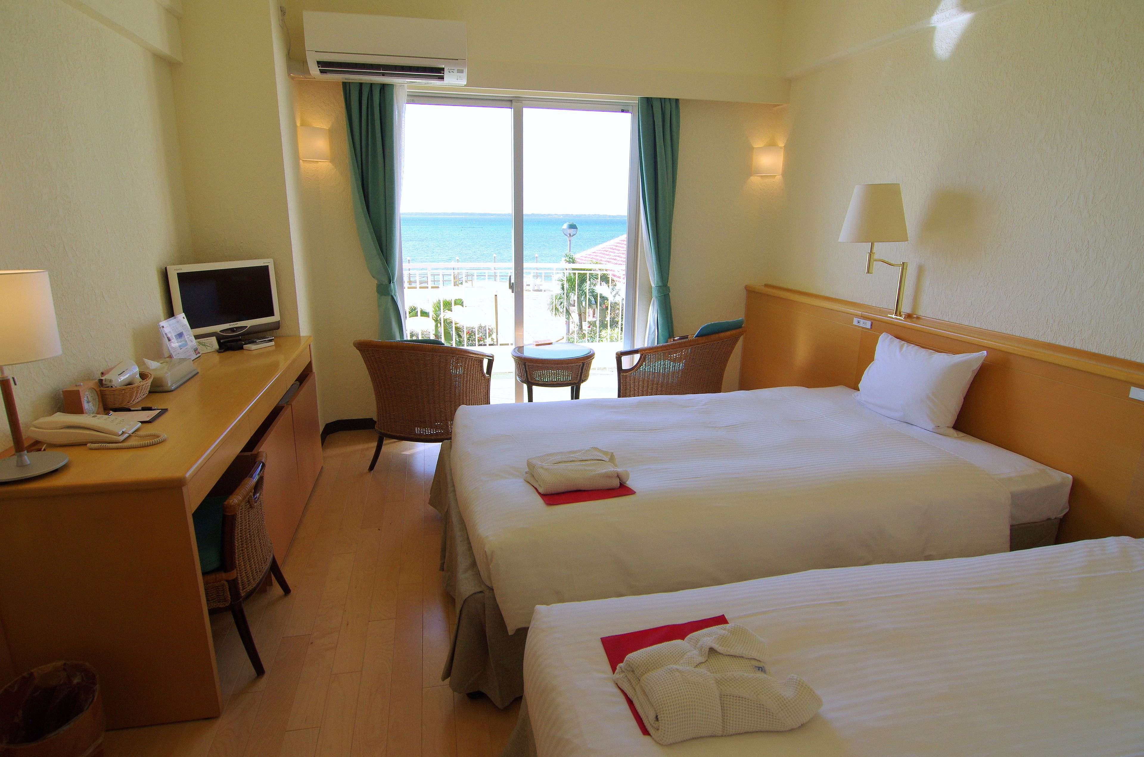 Habitación de hotel con dos camas, un escritorio con computadora, una silla, televisión y vista a la playa.