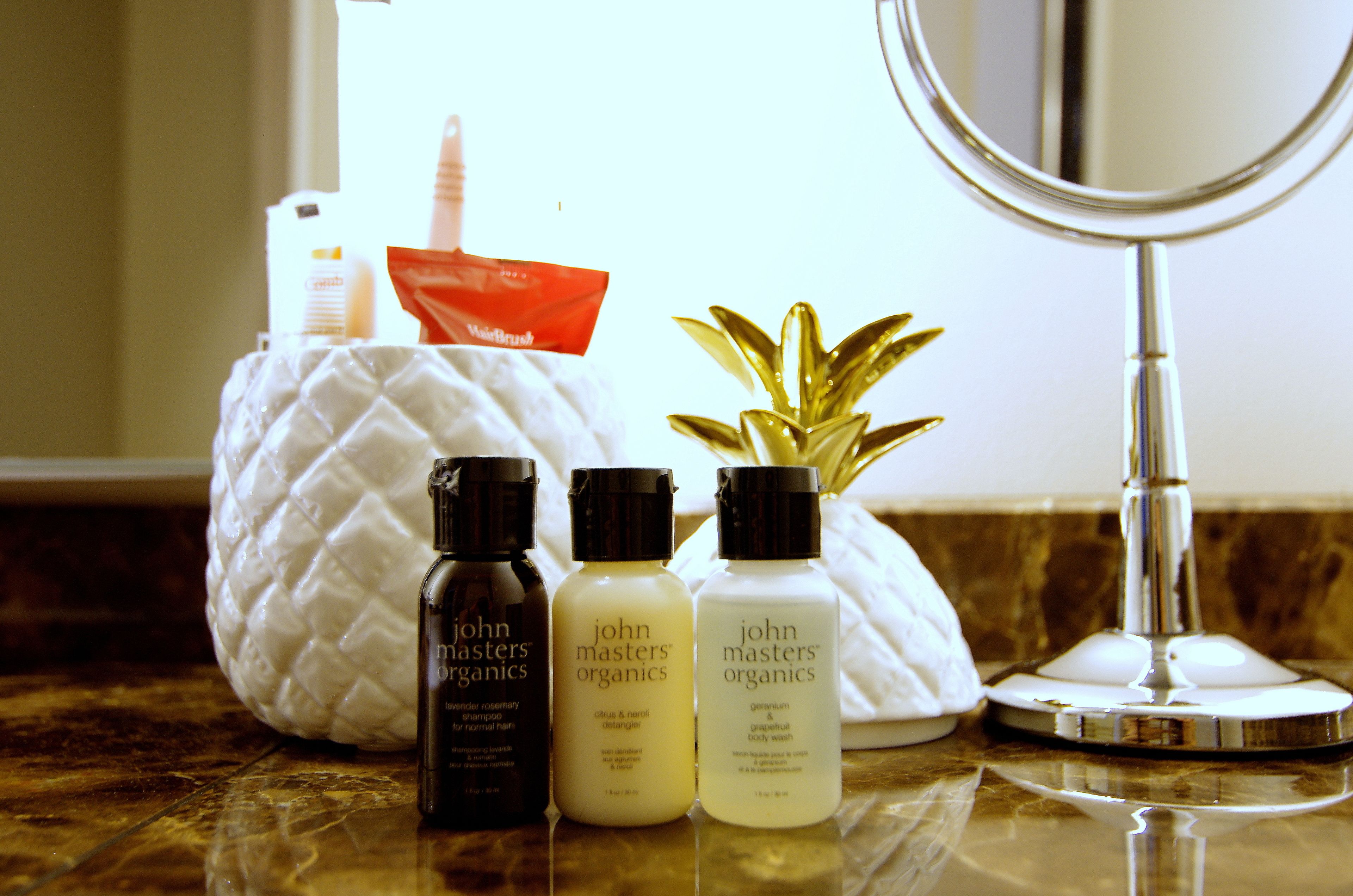 A bathroom counter with three bottles of John Masters Organics body wash, a red packet, and a potted plant.