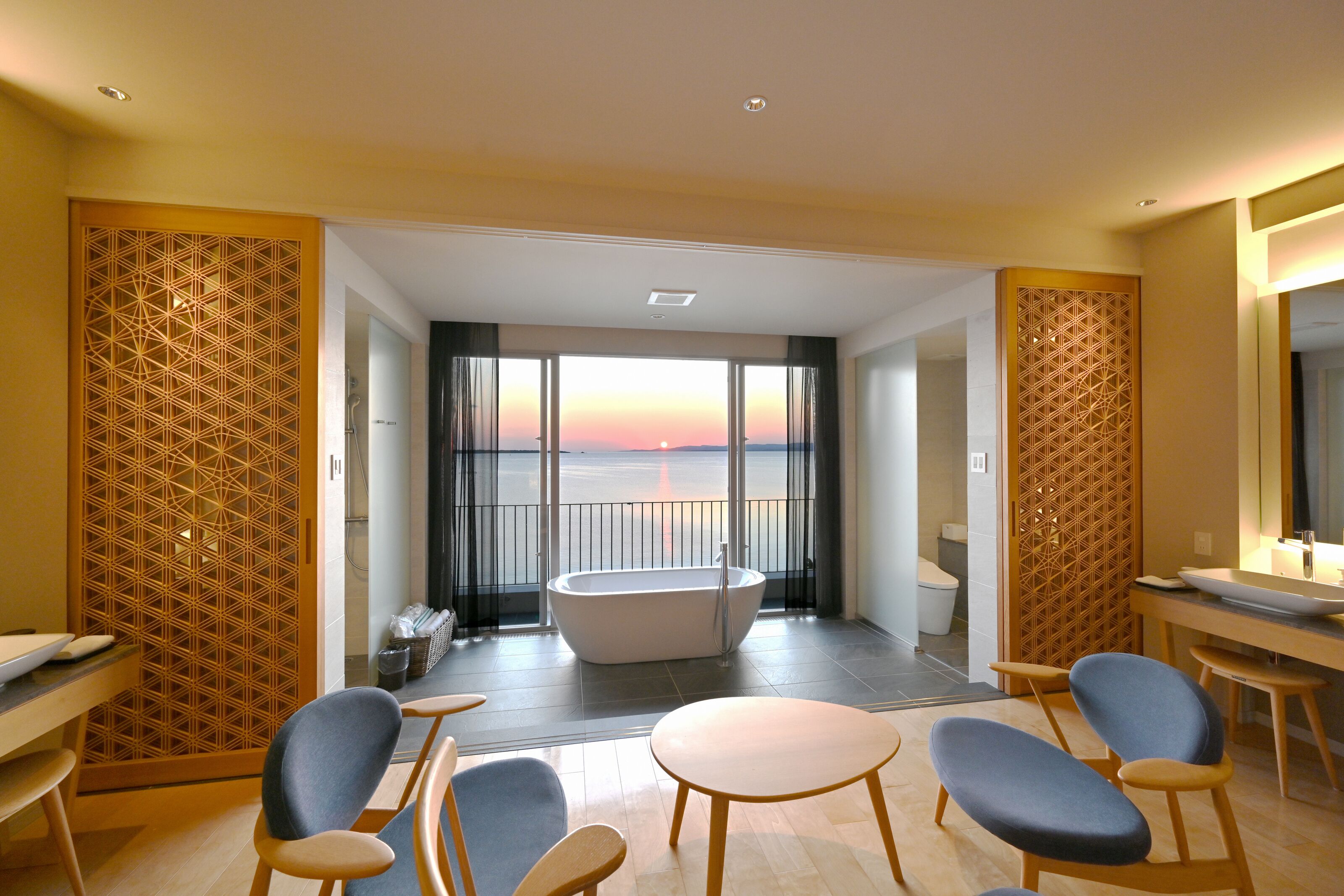 A modern hotel room with a large bathtub, a dining area with chairs, and a view of the sunset through large windows.