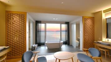 A modern hotel room with a large bathtub, a dining area with chairs, and a view of the sunset through large windows.