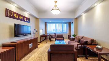 A hotel room with a flat-screen TV, a wooden entertainment unit, a sofa, a coffee table, and a large window with curtains.