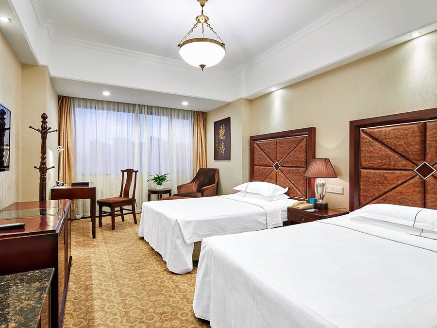 A hotel room with two beds, a desk, a chair, a TV, and a lamp.