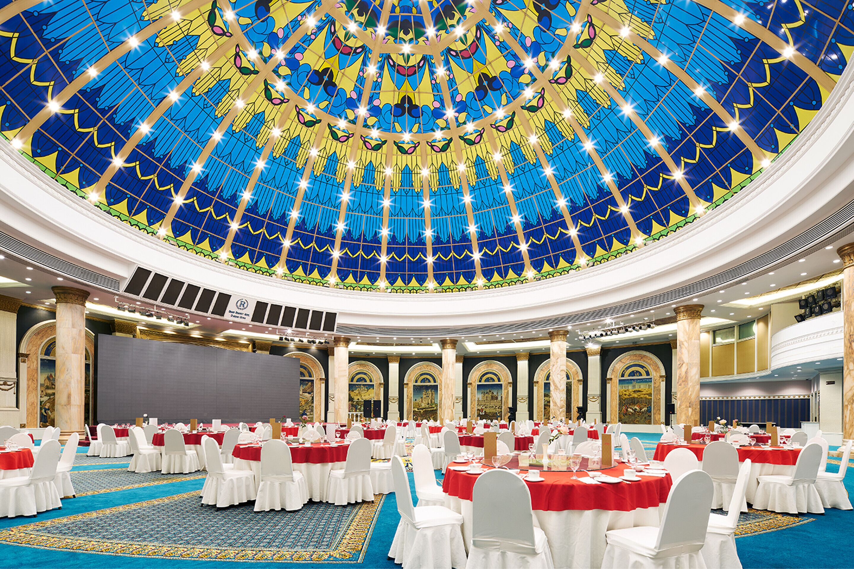 A large banquet hall with round tables, white chairs, and a blue carpet.
