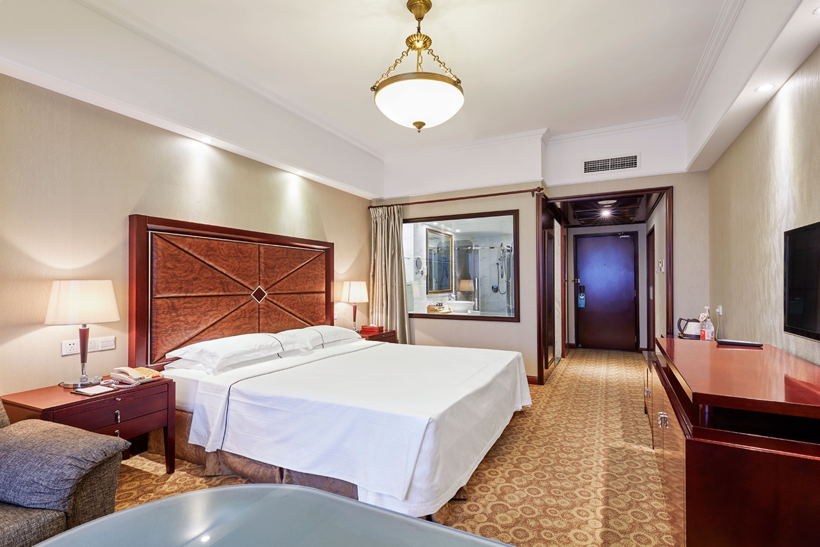 Quarto de hotel com uma cama grande, uma escrivaninha, uma cadeira, uma televisão e um banheiro visível através de uma porta de vidro.