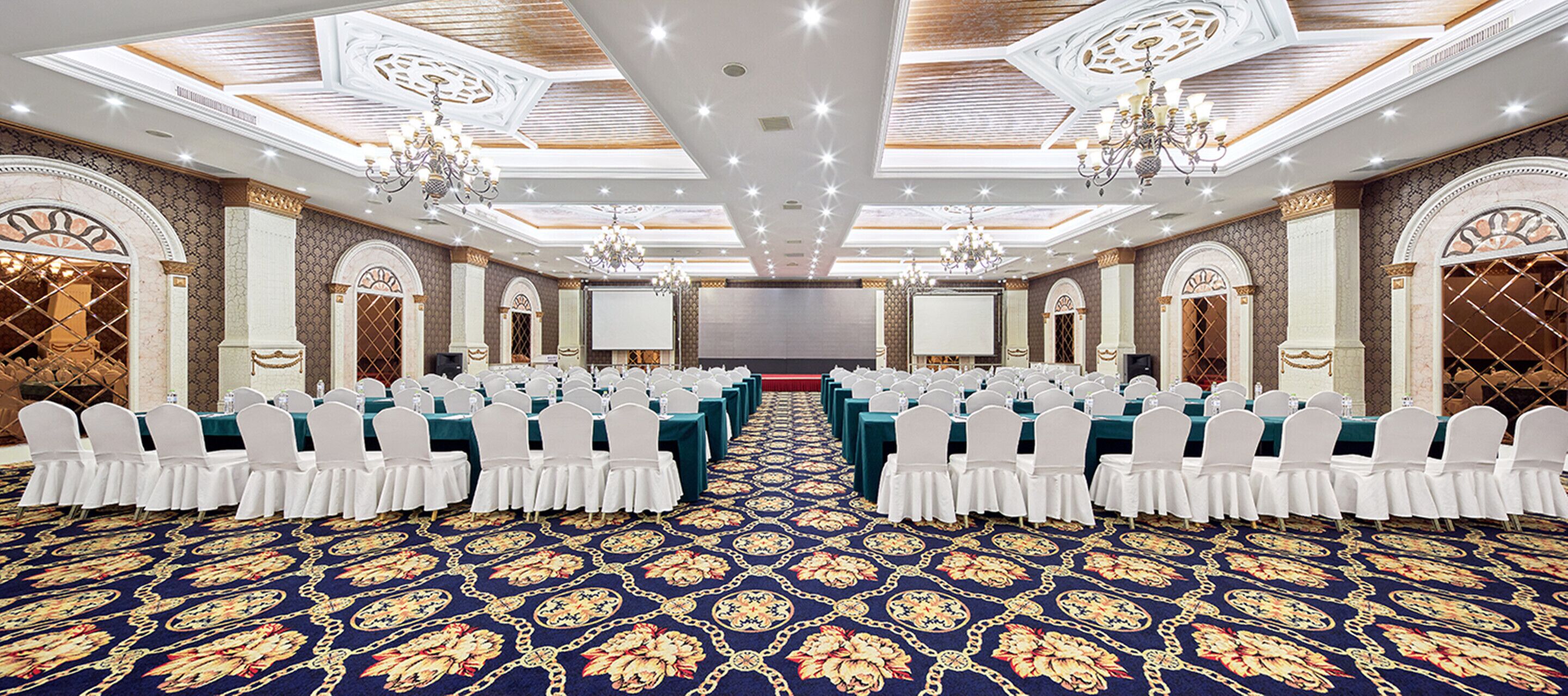 A conference room with rows of white chairs, a patterned carpet, and chandeliers.