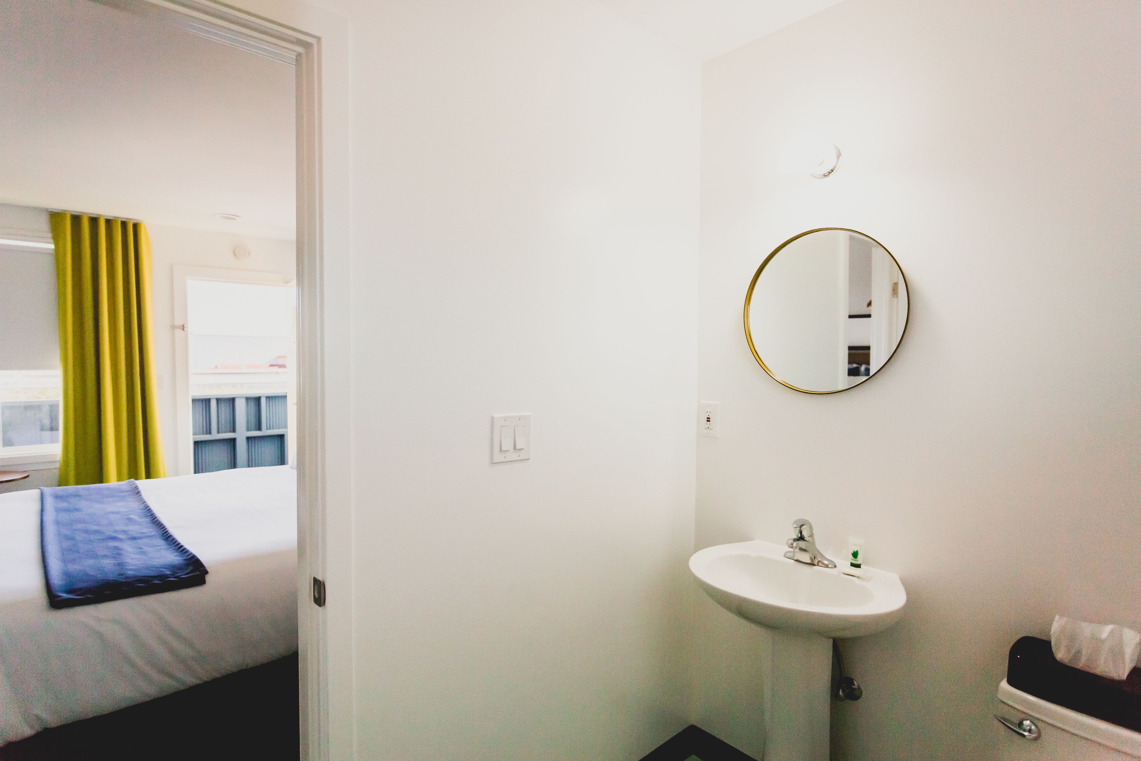 A hotel room with a bed, a white sink, a mirror, and a window with curtains.