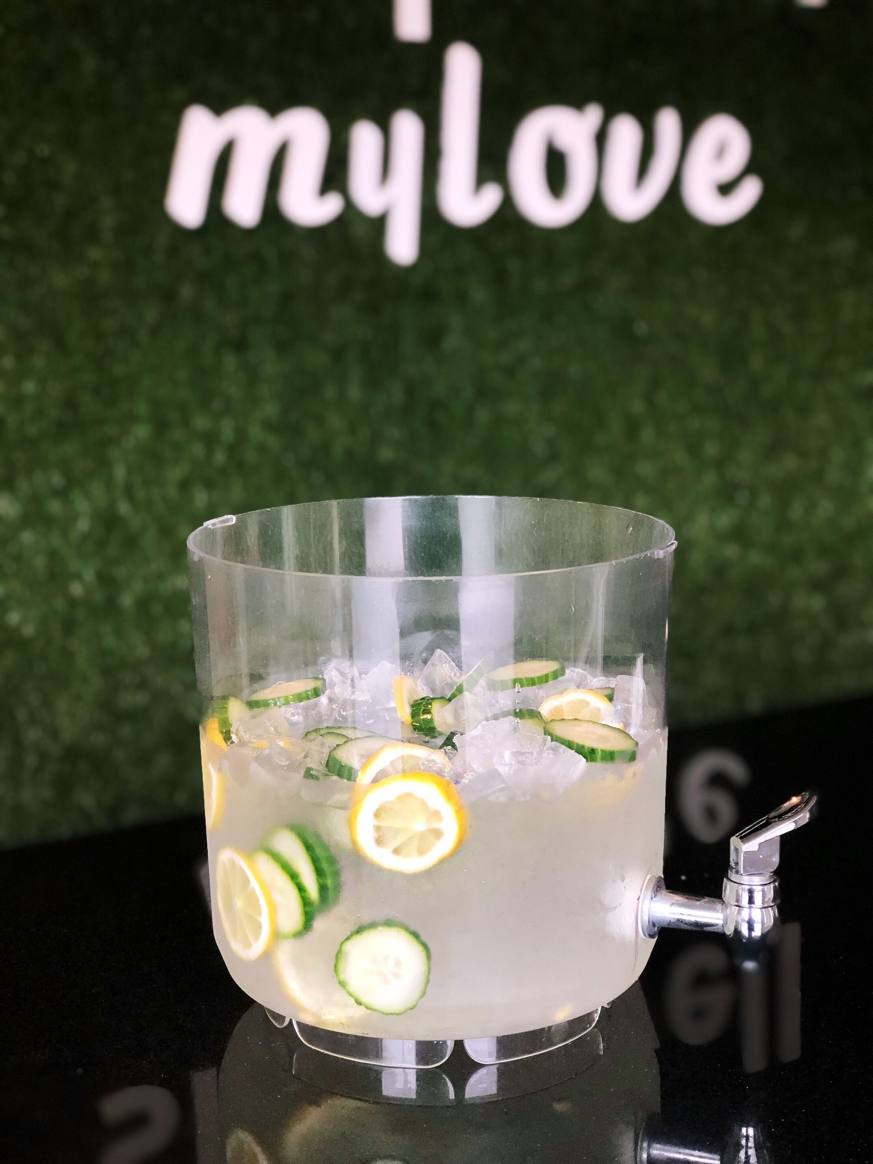 A clear glass with ice and lemon slices, placed on a reflective surface with a faucet.