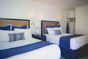 Two beds with white and blue bedding, a bedside table with a lamp, and a wall-mounted coat rack.