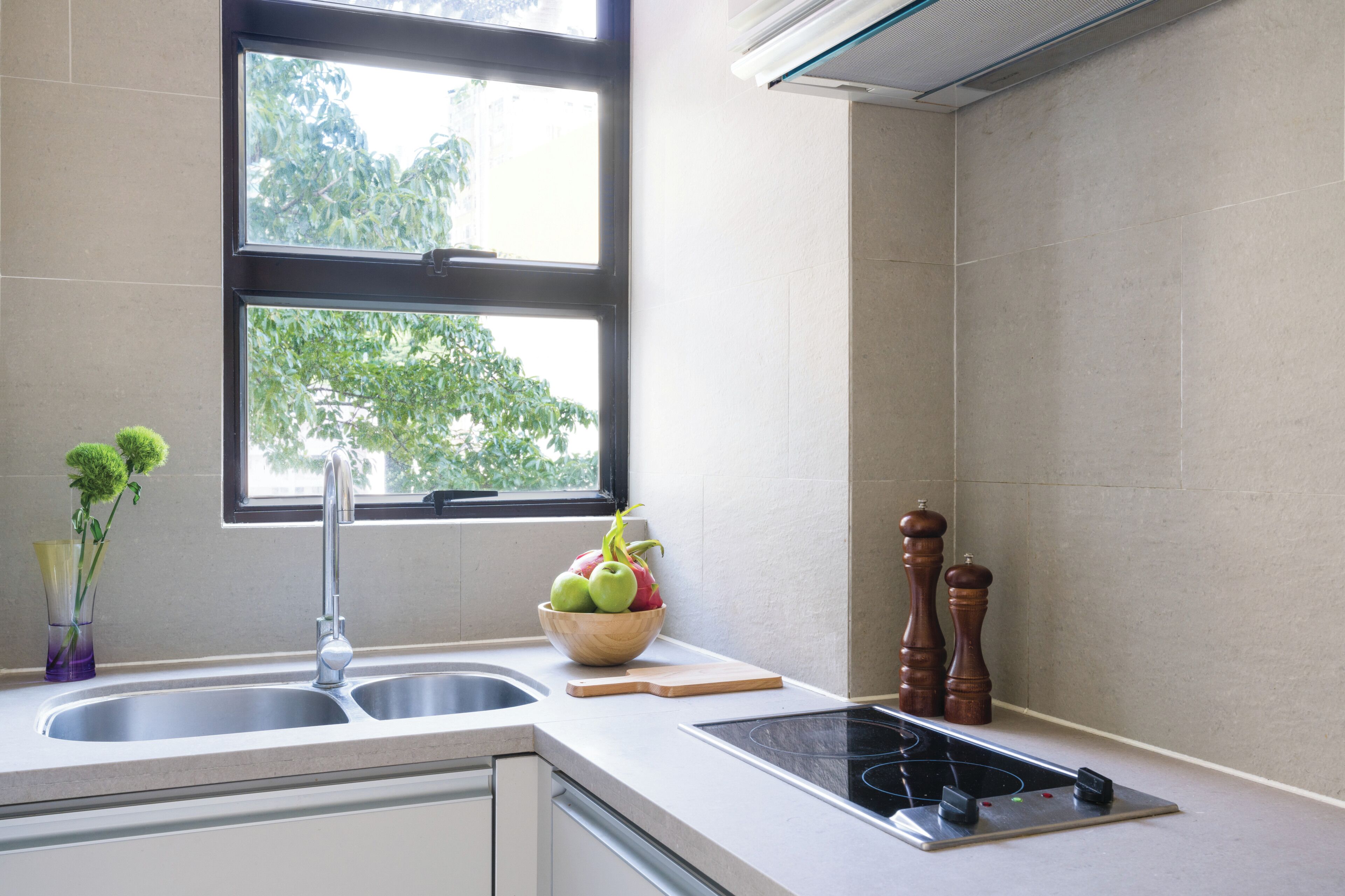 A kitchen with a double sink, a window, a bowl of fruit, a cutting board, a stove, and two wooden pepper shakers.