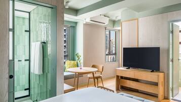 A modern hotel room with a bed, a TV, a small dining area, and a view of the city.