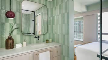 Un baño con pared de azulejos verdes, un lavabo blanco, un espejo y una mesita de noche junto a la cama.