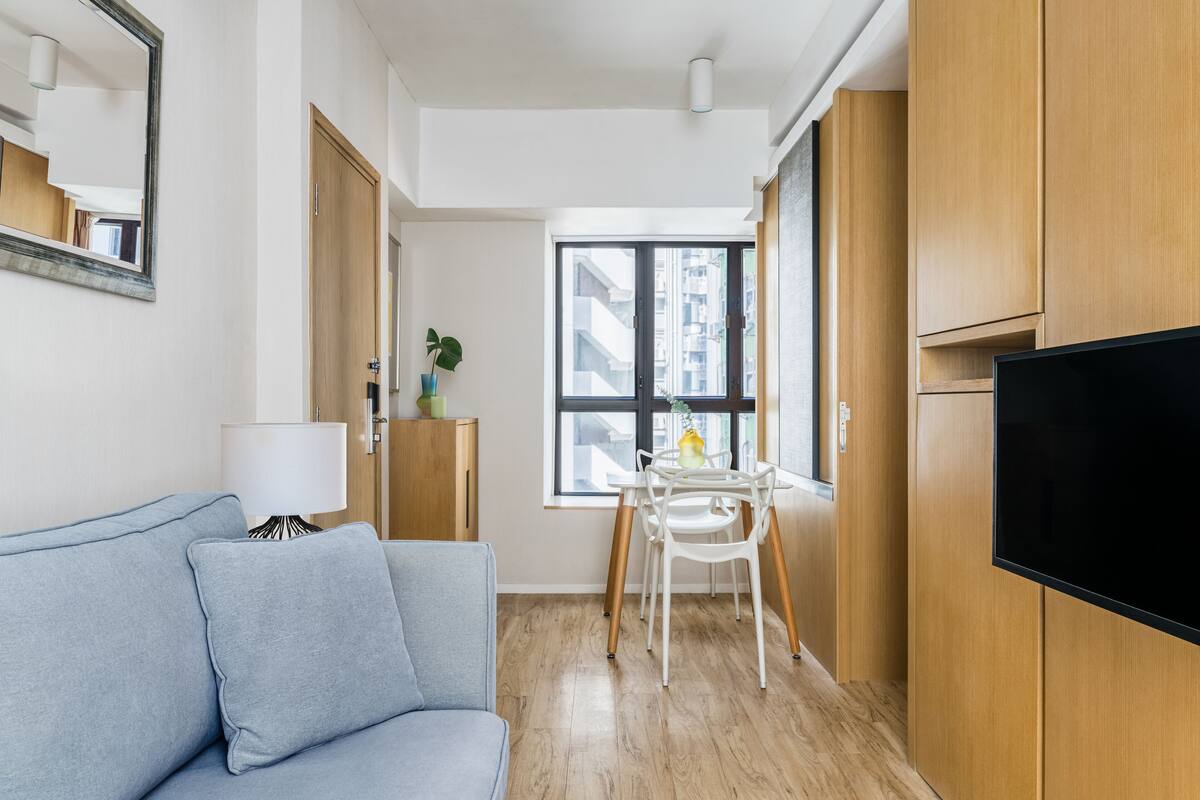 A modern living room with a sofa, a TV, a dining table with chairs, and a window with a view of buildings.