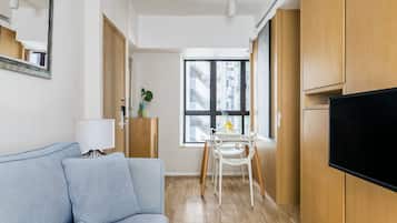 A modern living room with a sofa, a TV, a dining table with chairs, and a window with a view of buildings.