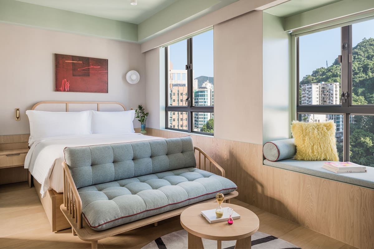 A modern hotel room with a bed, a sofa, a round table, and a window with a view of buildings.