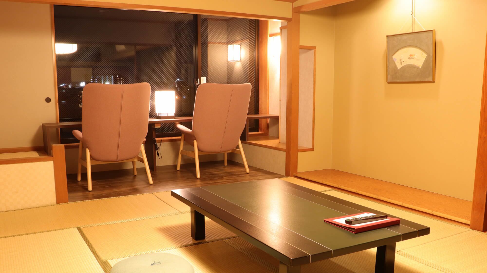Ein Zimmer mit Tatami-Boden, einem niedrigen Tisch, zwei Stühlen, einer Schiebetür und einem gerahmten Bild an der Wand.